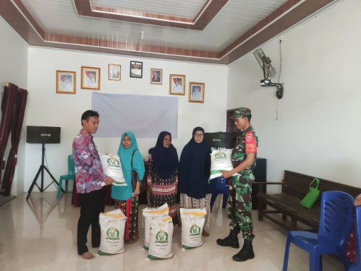 Seorang Bintara Pembina Desa Sidrap menyerahkan karung beras bantuan pangan kepada warga penerima manfaat di sebuah ruangan penyaluran bantuan, disaksikan petugas pemerintah setempat dan beberapa penerima.
